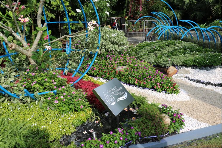 A colorful garden with flowers, green plants, blue sculptures, and a sign reading "Gardens by the Bay" near a gravel path.